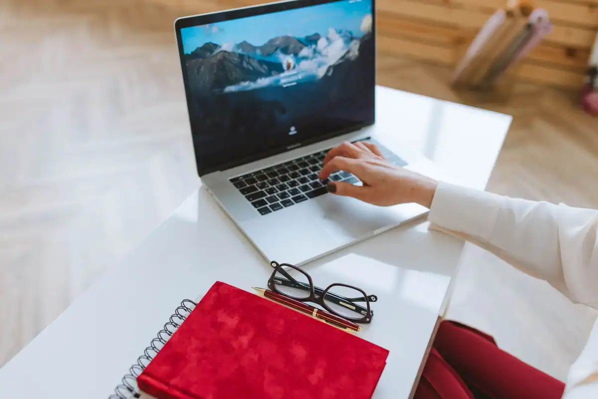 Persoană care lucrează la laptop, cu un carnet roșu și ochelari pe o masă albă
