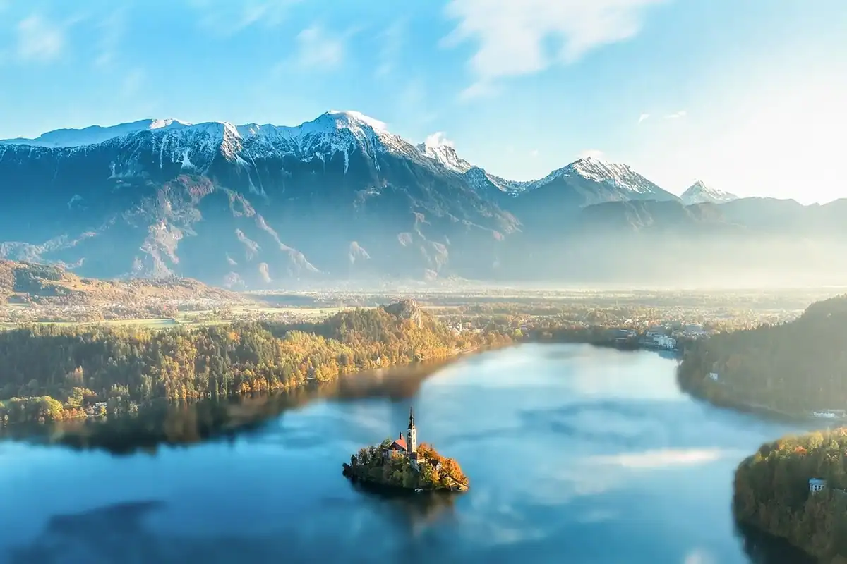 Lac de munte cu apă liniștită, văzut de sus, cu ceață deasupra și munți în fundal