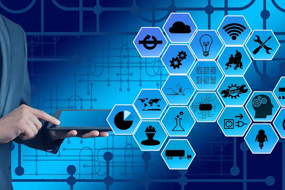 Mână cu tabletă în fața unor pictograme hexagonale pentru dispozitive conectate