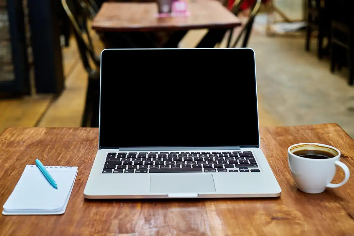 Laptop deschis pe o masă de cafenea, lângă o ceașcă și un carnet