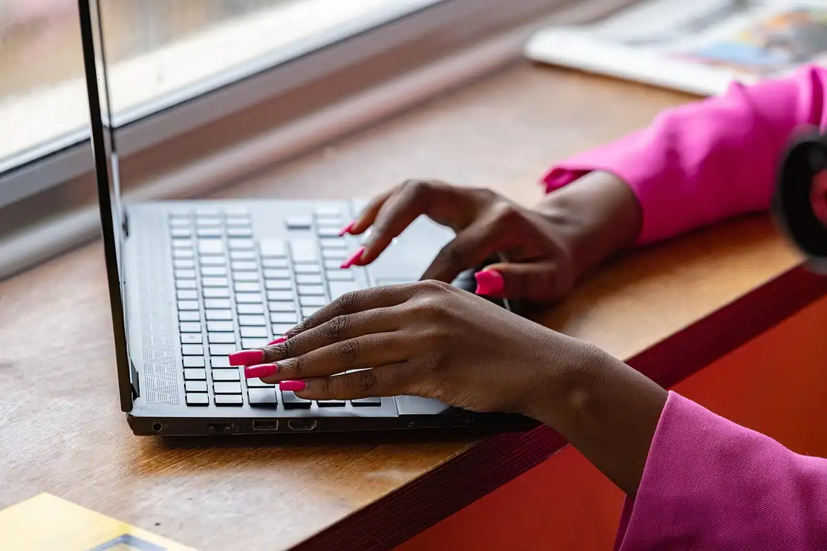 Femeie cu mânecă roz care tastează pe un laptop lângă fereastră